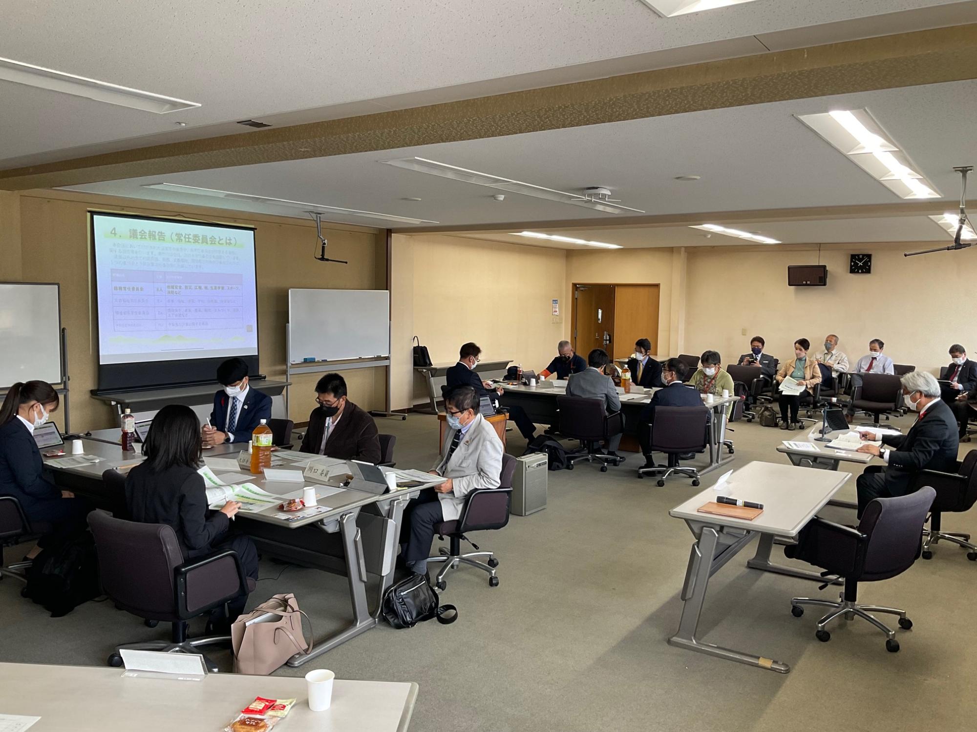 前方のスクリーンに資料が映し出される中、人々がテーブルを囲んで着席し、手元の資料やノートパソコンを見ている総務常任委員会の様子の写真