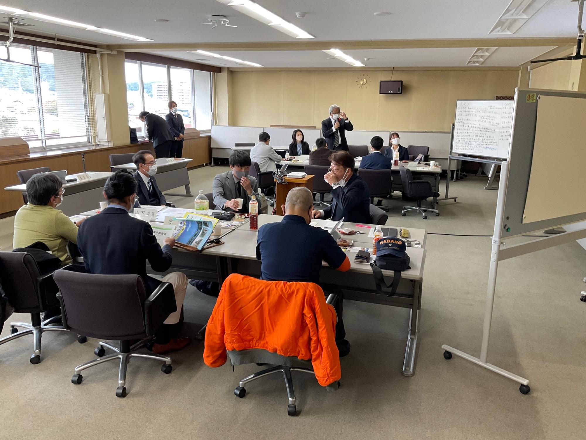 複数のグループがテーブルを囲んで着席し、資料を見ながら議論しており、奥では男性が話し、ホワイトボードに書き込みがある様子の写真