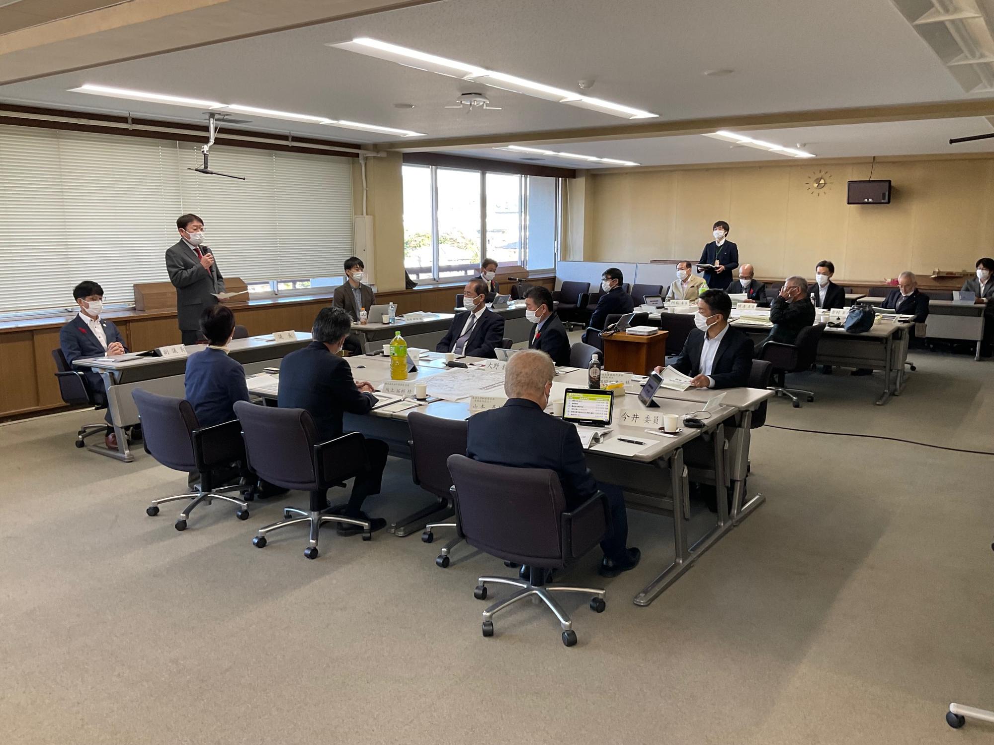 明るい会議室で、多くのスーツ姿の参加者がテーブルに着席し、左手前で立つ男性がマイクで発表しているのを、聞いている真剣な会議の様子の写真