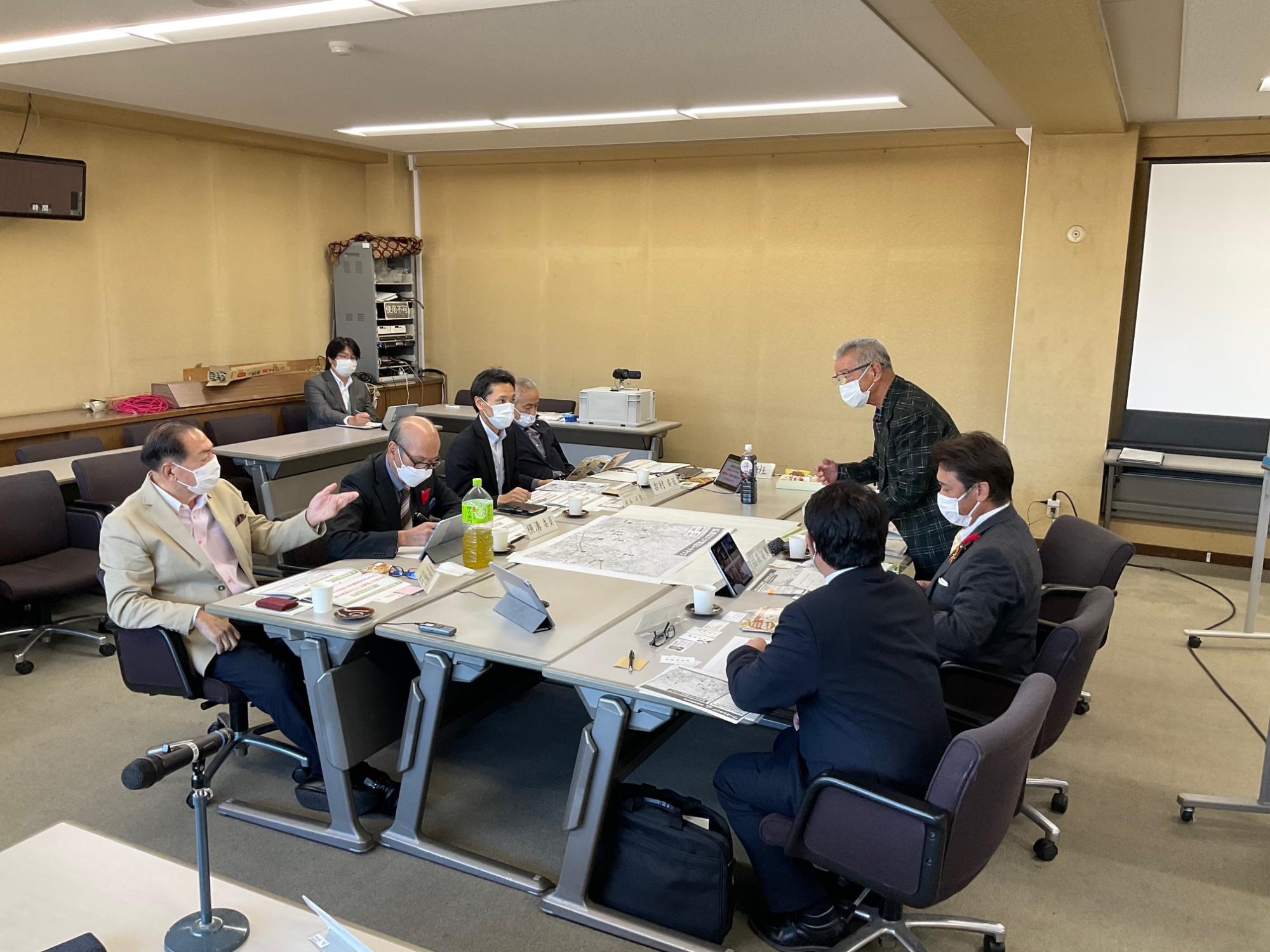 会議室で、大きなテーブルに地図のような資料を広げ、マスク姿の男性たちが議論しており、一人が身振りを交えて話し、別の男性が立ってテーブルを覗き込んでいる様子の写真