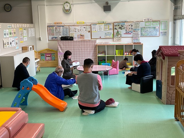 緑のマットが敷かれた部屋で、滑り台やおもちゃが置かれる中、スーツ姿の男性を含む複数の大人が床に座り込み、資料を見ながら話し合っている様子の写真