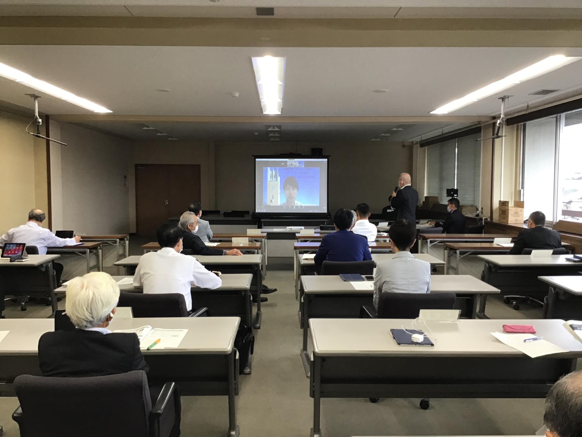 部屋の前方に設置された、講師の男性が写ったスクリーンの方を向いて受講者たちが席に座り、1人の男性が自席から起立し、スクリーンの方を向いて講師の男性と会話をしている様子の写真