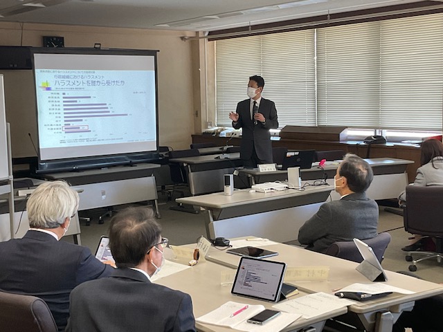 会議室で棒グラフのデータが映し出されたスクリーンの右側に立った講師の男性が、席に座った聴講者へ説明をしている様子の写真