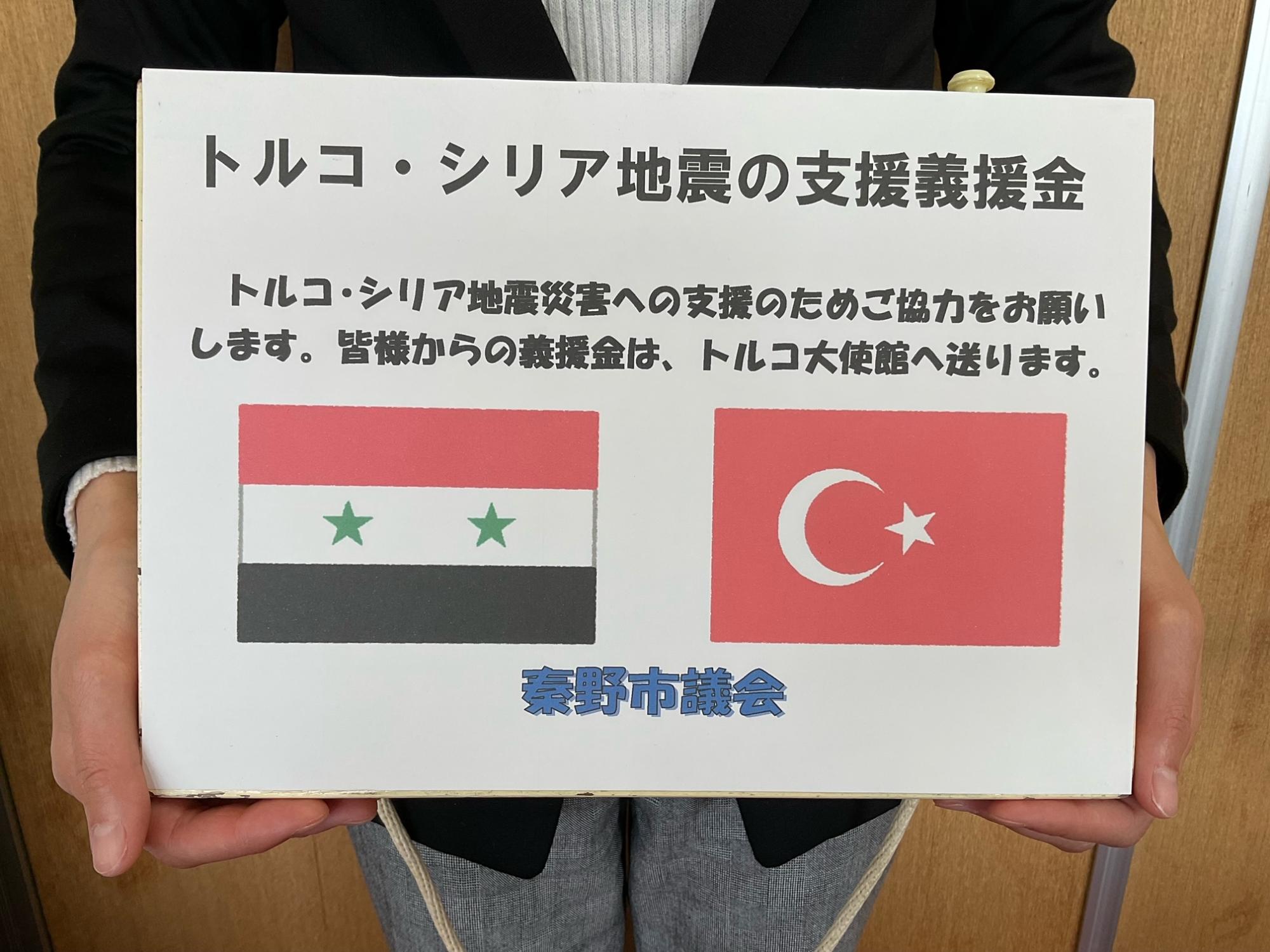 トルコ・シリア地震の支援義援金を呼びかけるプラカードの写真で、トルコの国旗とシリアの国旗が並び「秦野市議会」と印字されている写真