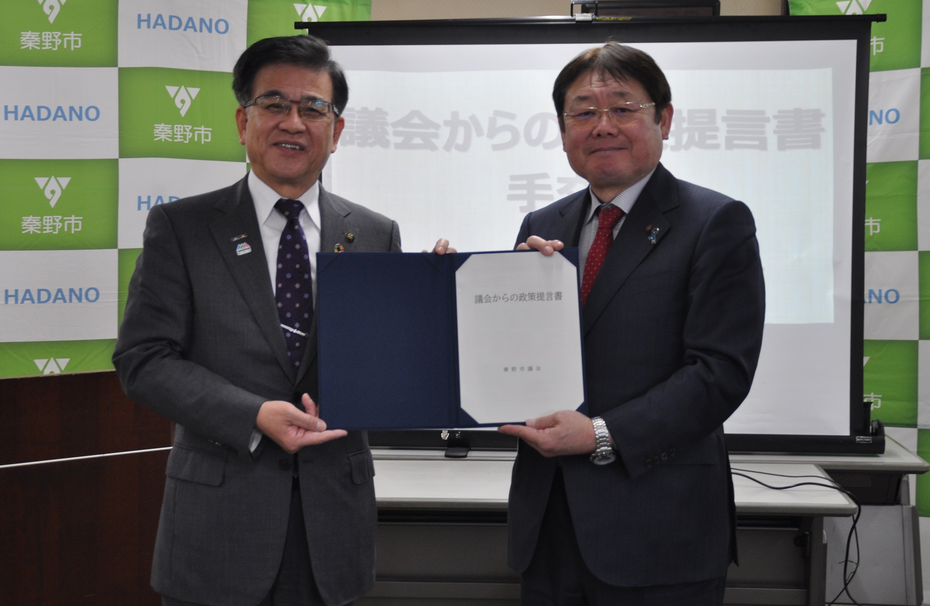 「秦野市」のロゴが並ぶ背景の前で、スーツ姿の議長と市長が「議会からの政策提言書」と記された青いファイルを開いてカメラに向け、笑顔で並んでいる写真