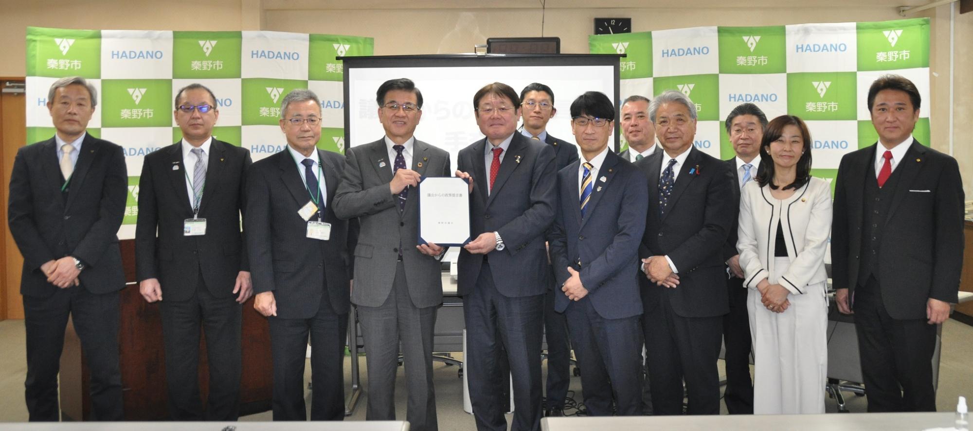 「秦野市」のロゴがある背景の前で、スーツ姿の男女12人が並び、中央の市長と議長が青いファイルに挟まれた書類を一緒に持ってカメラを見ている集合写真