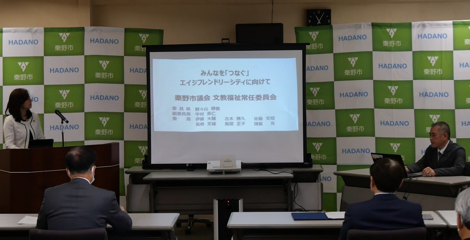 「秦野市」ロゴを背景に、「みんなを「つなぐ」エイジフレンドリーシティに向けて」と題したスライドが映る前で、演台の女性が発表し、着席した数名が聞いている文教福祉プレゼンの様子の写真