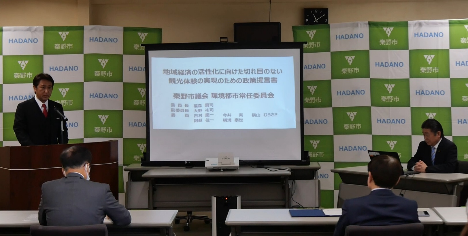 「秦野市」のロゴが並ぶ背景の前で、スーツの男性が演台に立ち、スライドを背に発表している環境都市プレゼンの様子の写真