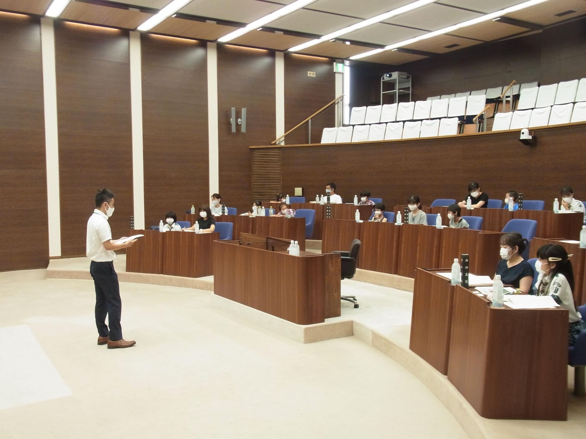 マスクを着用した男性が説明しているのを、議席には子どもと保護者らが着席して真剣に聞いている様子の写真