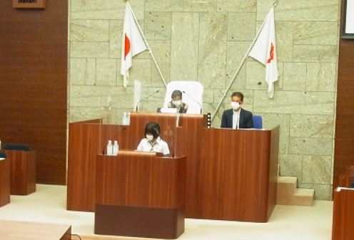 日本の国旗が飾られた壇上にマスクをつけた大人たちが座り、一人の子供が演台に立ちマイクに向かって何かを発言している様子の写真