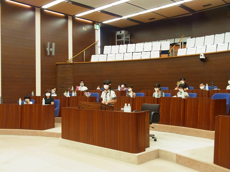 木製の議場で、小学生の女の子が演説台に立って発表しており、他の多くの子供たちがその様子を座って聞いている写真