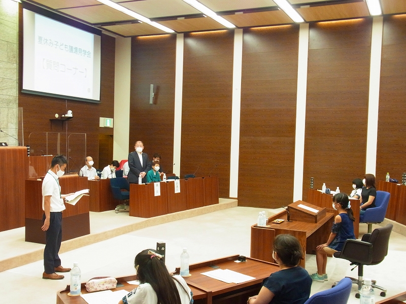 「夏休み子ども議場見学会【質問コーナー】」と書かれたスクリーンを背景に、着席した子どもが質問し、壇上の大人たちが答えている様子の写真