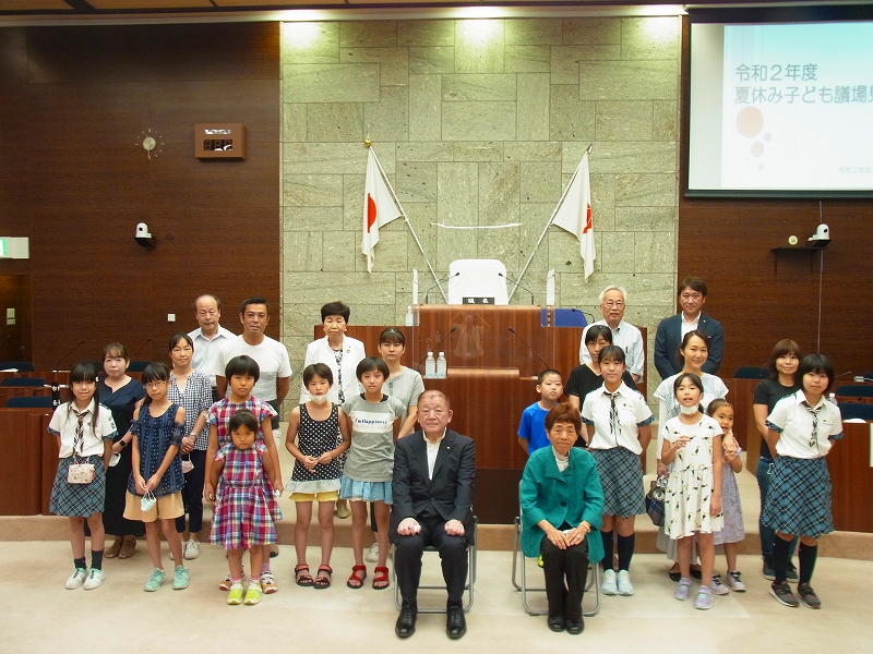 木目調の壁と日本の国旗が飾られた議場で、スーツ姿の正副議長が椅子に座り、子供たちと一緒に立って記念撮影をしている「夏休み子ども議場見学」イベントの集合写真