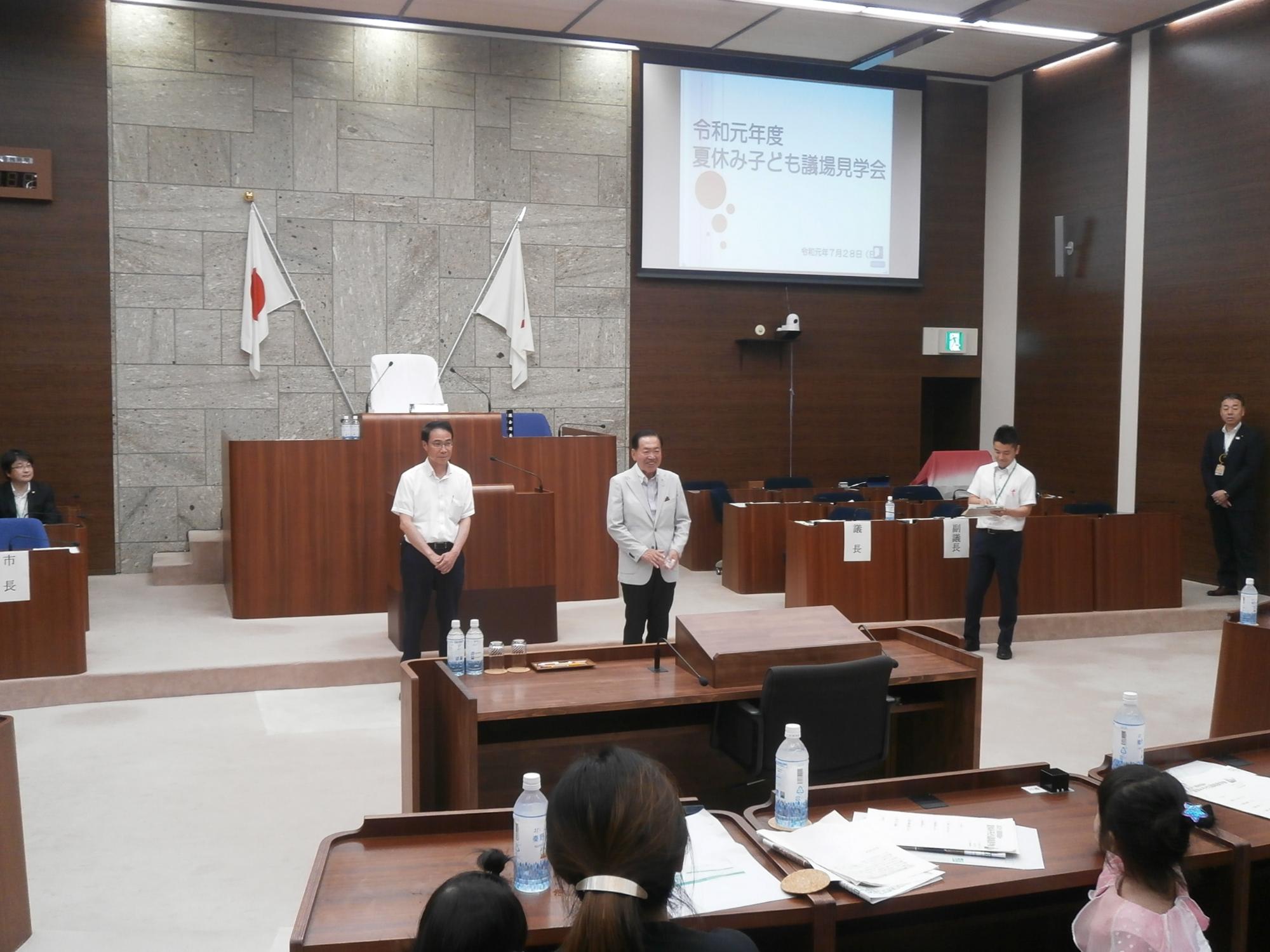 白いスクリーンの前に立つ男性たちと、それを机に座って見つめる多くの親子が写っている、議会見学会の様子の写真