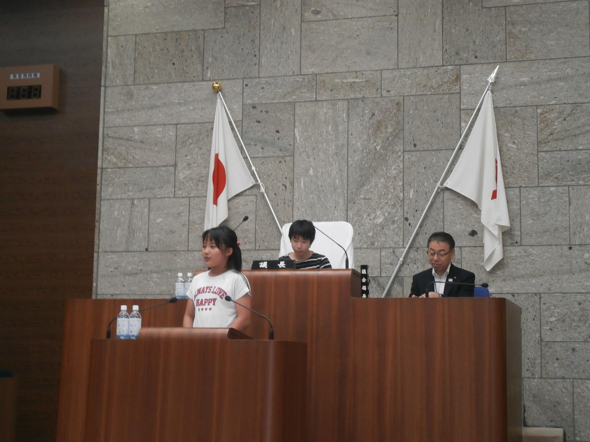 日本の国旗が飾られた壇上で、女の子がマイクで発言しており、後ろに座る大人たちが静かに耳を傾けている、子ども議会の様子の写真