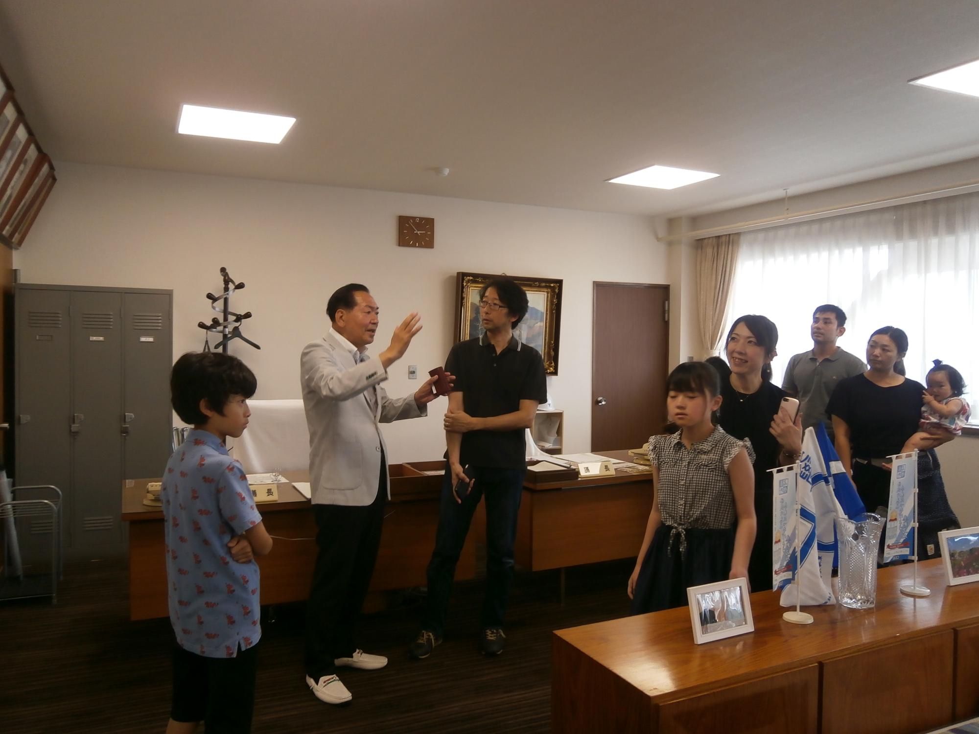 スーツ姿の議長が訪れた子供たちと大人に身振り手振りを交えながら話をしており、和やかな雰囲気で交流が行われている様子の写真