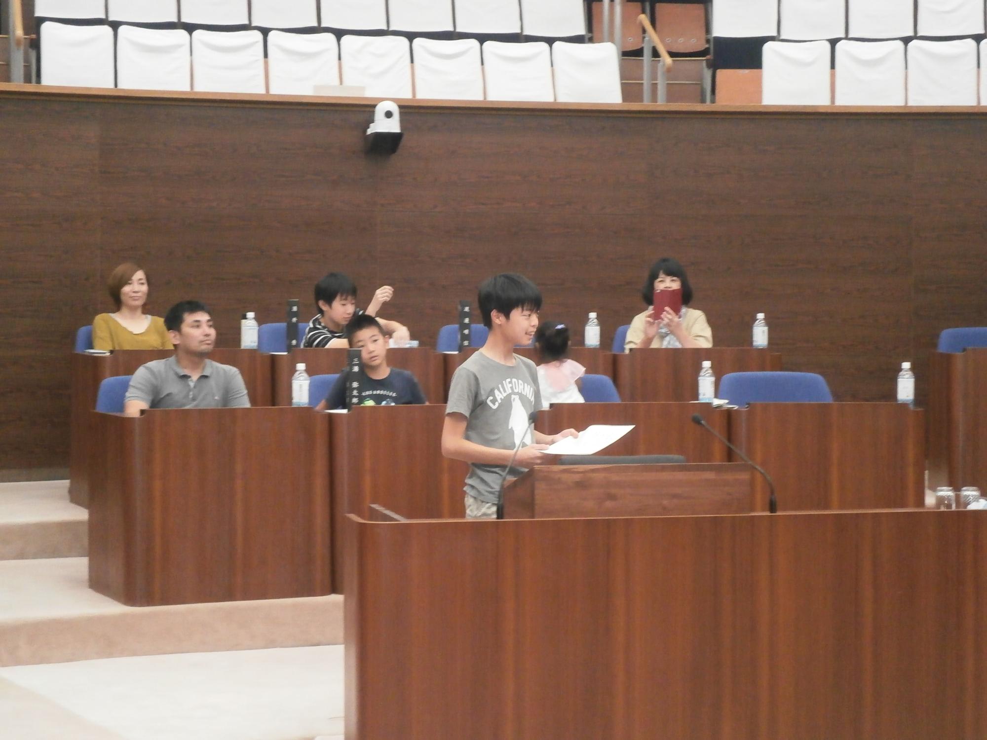 男の子が演説台に立ってマイクで発言しており、他の子供や大人たちがその様子を静かに見守っている、子ども議会の様子の写真