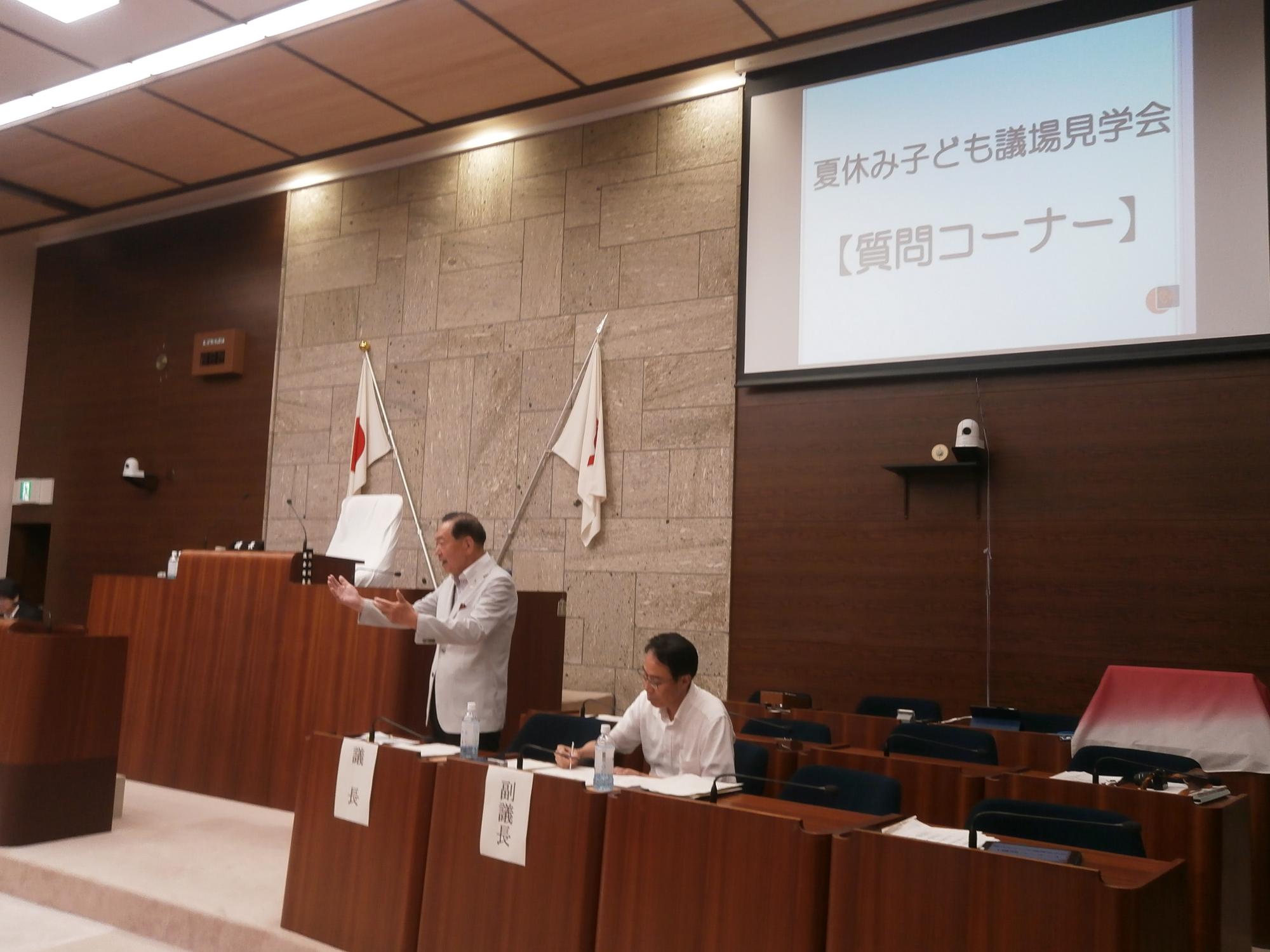 「夏休み子ども議場見学会【質問コーナー】」と映し出されたスクリーンを背景に、スーツ姿の議長がジェスチャーを交えながら回答している様子の写真