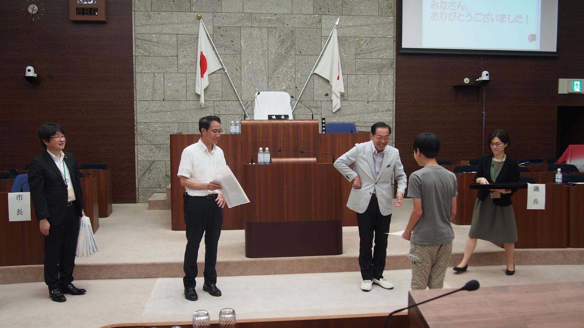 日本の国旗がある議場で、壇上の大人たちが子供たちに修了証を手渡し、和やかな雰囲気の中で行われている見学イベントの様子の写真