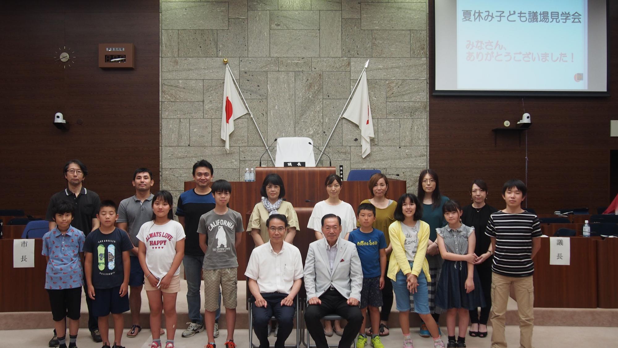 大理石と木目調の壁の議場で、日本の国旗を背景に「夏休み子ども議場見学会」と書かれたスクリーンがあり、スーツを着た議長と多くの子供たちが和やかに記念撮影をしている写真