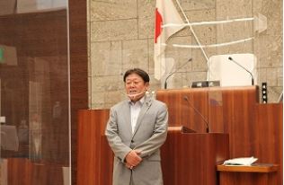 神奈川県秦野市の議会本会議場で、スーツ姿の小菅議長が透明なアクリル板の前で立って話している様子が写っている写真