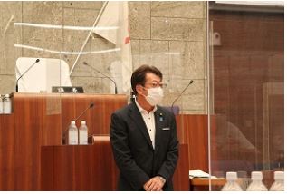 神奈川県秦野市の議会本会議場で、黒いスーツと白いマスクを着用した谷副議長が透明なアクリル板の前で立っている様子が写っている写真