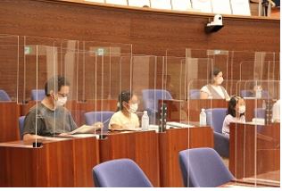 パーテーションで仕切られた議場で、マスクを着用した子供たちと大人が着席しており、何かを熱心に読んだり書き込んだりしている様子の写真