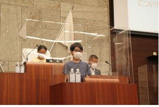 マスクをつけた男の子が壇上に立ち、その横にはマスクをつけた大人たちが座って見守っている、議会体験の様子の写真