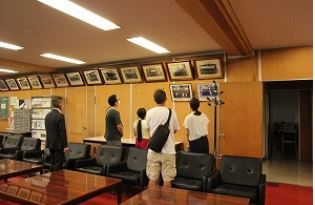 スーツ姿の男性が数人の見学者を案内し、大人と子どもが壁に飾られた歴代市長の写真を見上げている様子が写っている写真