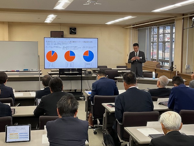 講師の男性が円グラフが表示された大型スクリーンの右側に立ち、前方を向いて席に座った聴講者へ話をしている研修の様子の写真