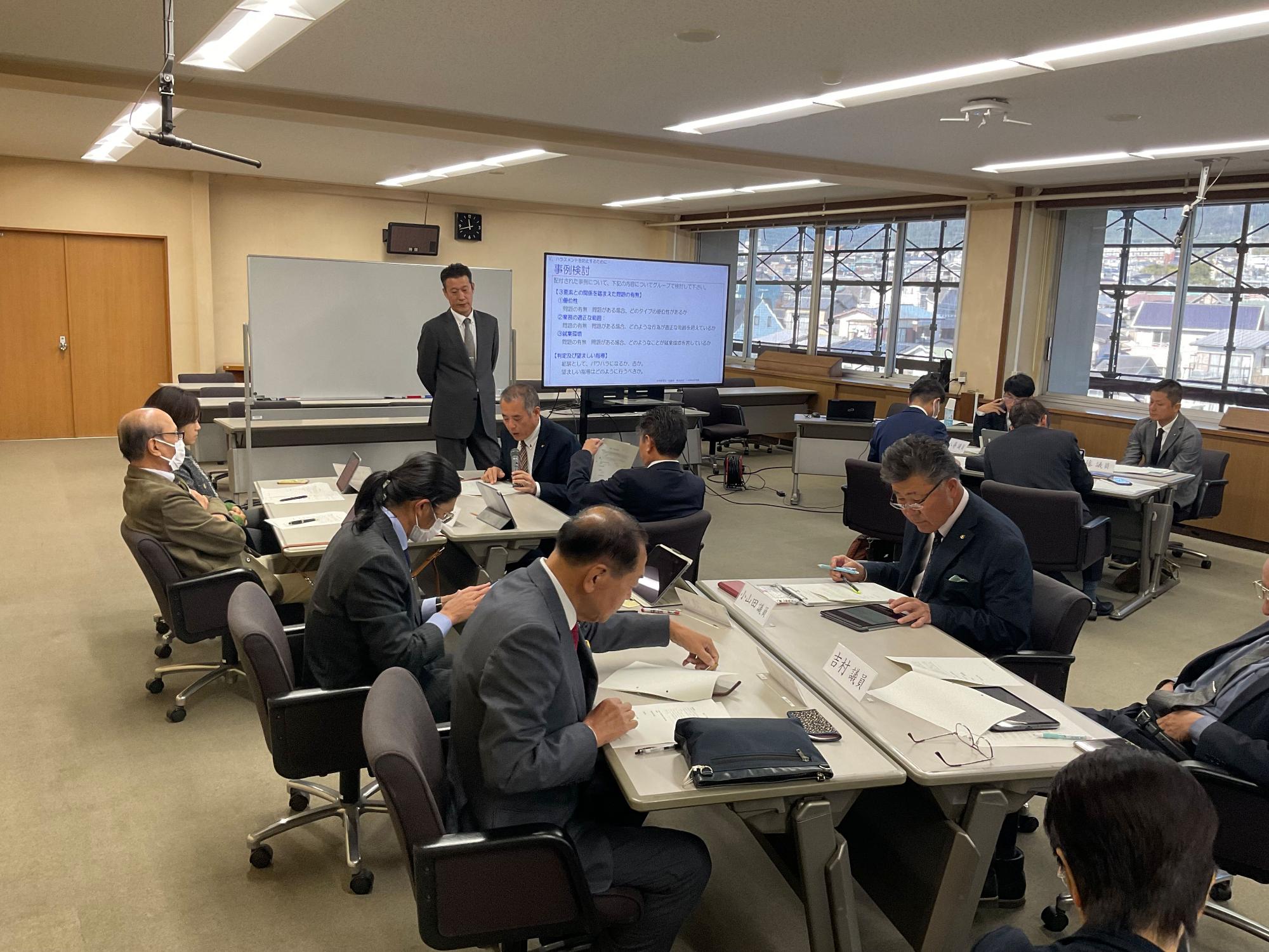 大きな窓がある会議室で、スーツ姿のたくさんの男性が長机を囲んで資料を見ながら話し合いを行なっており、講師の男性がホワイトボードを背にして立って監督している様子の写真