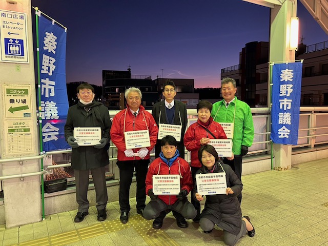 駅のホームで、秦野市議会と書かれた青いのぼりの下に、赤いベストや緑のジャンパーを着て募金箱と「令和6年能登半島地震災害支援」のプラカードを持った7人の男女の集合写真