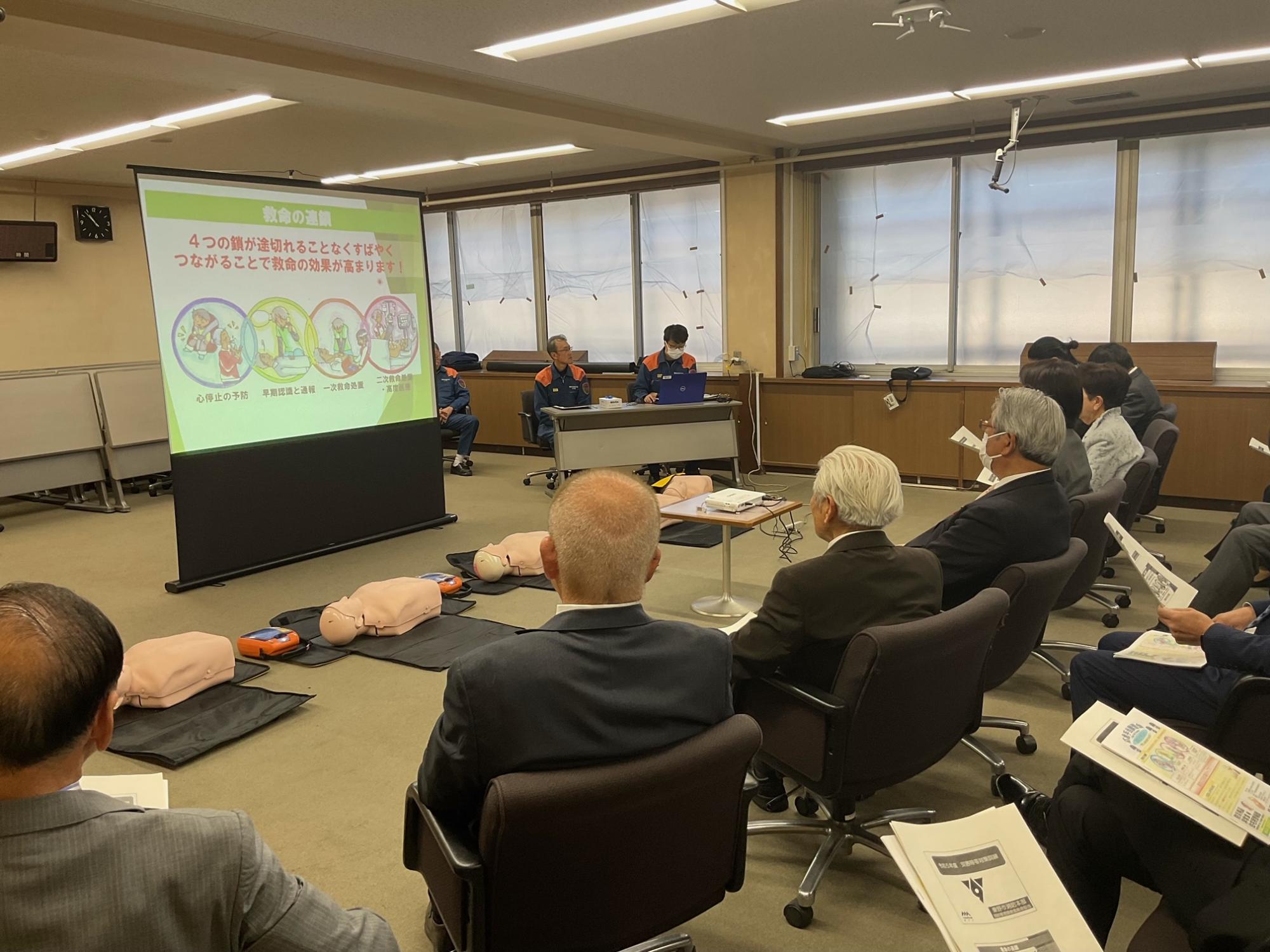 大会議室のような場所で、スーツ姿の参加者たちが消防職員たちから、プロジェクターに映し出されたスライドと床に置かれた人形やAEDを使って応急手当の方法を学んでいる救命講習の風景写真