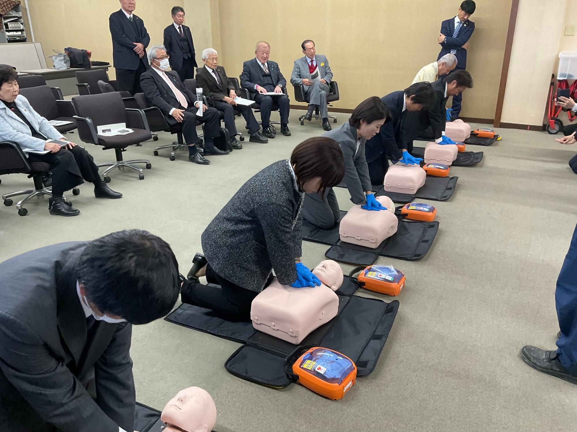 スーツ姿の参加者たちが消防職員から心肺蘇生法を教わり、床に置かれた等身大の人形を使って胸骨圧迫の実技訓練を行なっている様子の写真