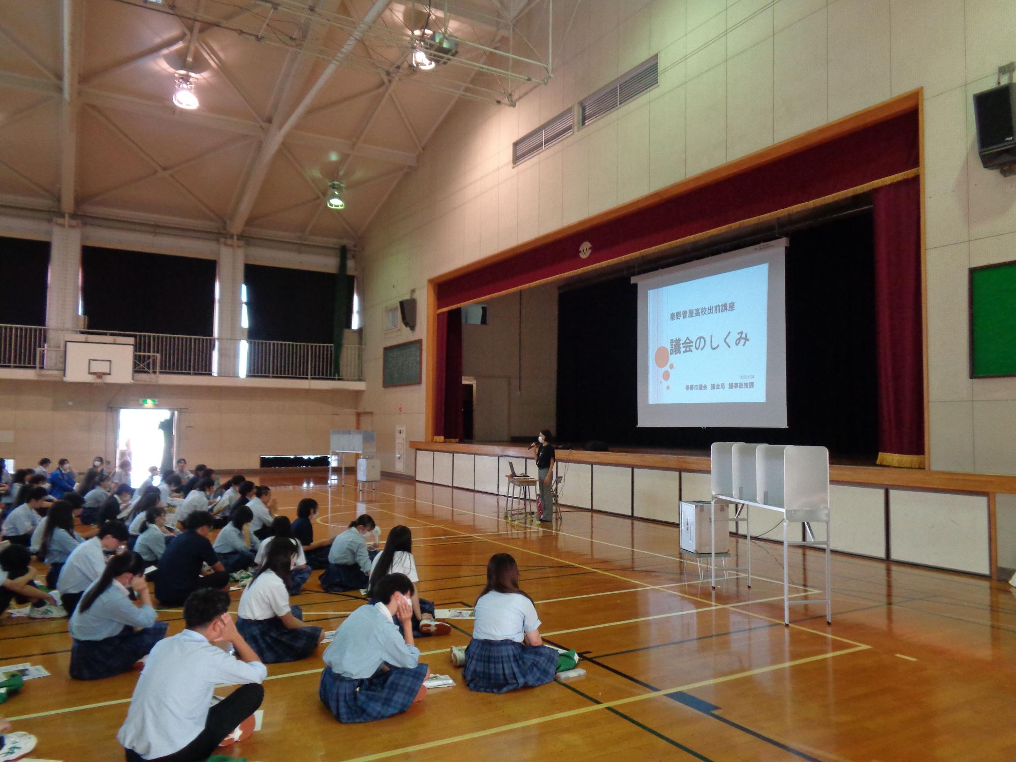 体育館の床に制服姿の生徒が大勢座り、壇上のスクリーンには「議会のしくみ」というタイトルの資料が投影され、一人の講師がマイクを持って立っている秦野曽屋高校出前講座の写真