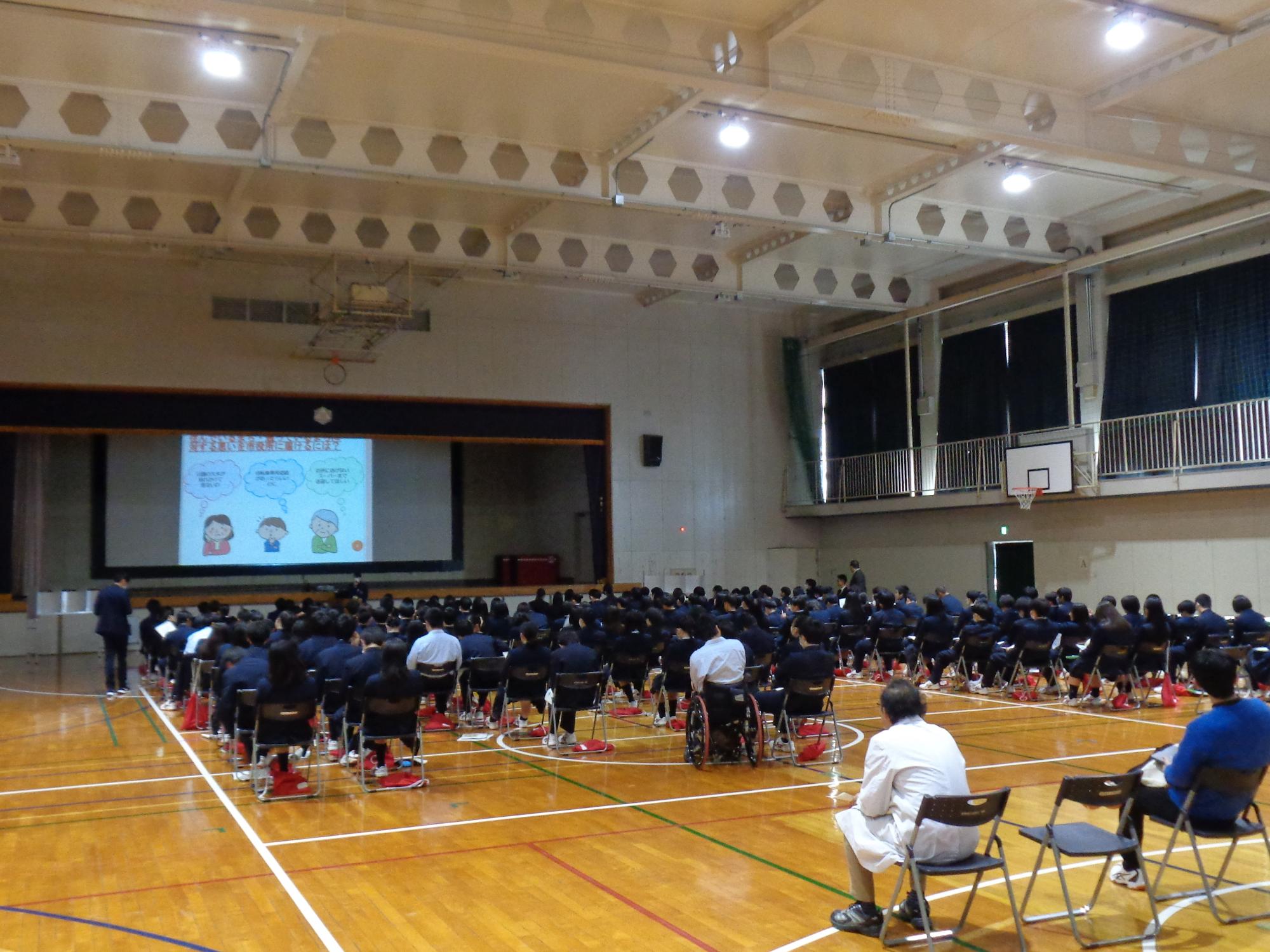 体育館の床に置かれた椅子に、黒い制服姿の生徒が大勢座って体育館中央の壇上にあるスクリーンを見ており、スクリーンに資料と3人の人物イラストが投影されている秦野総合高校出前講座の写真