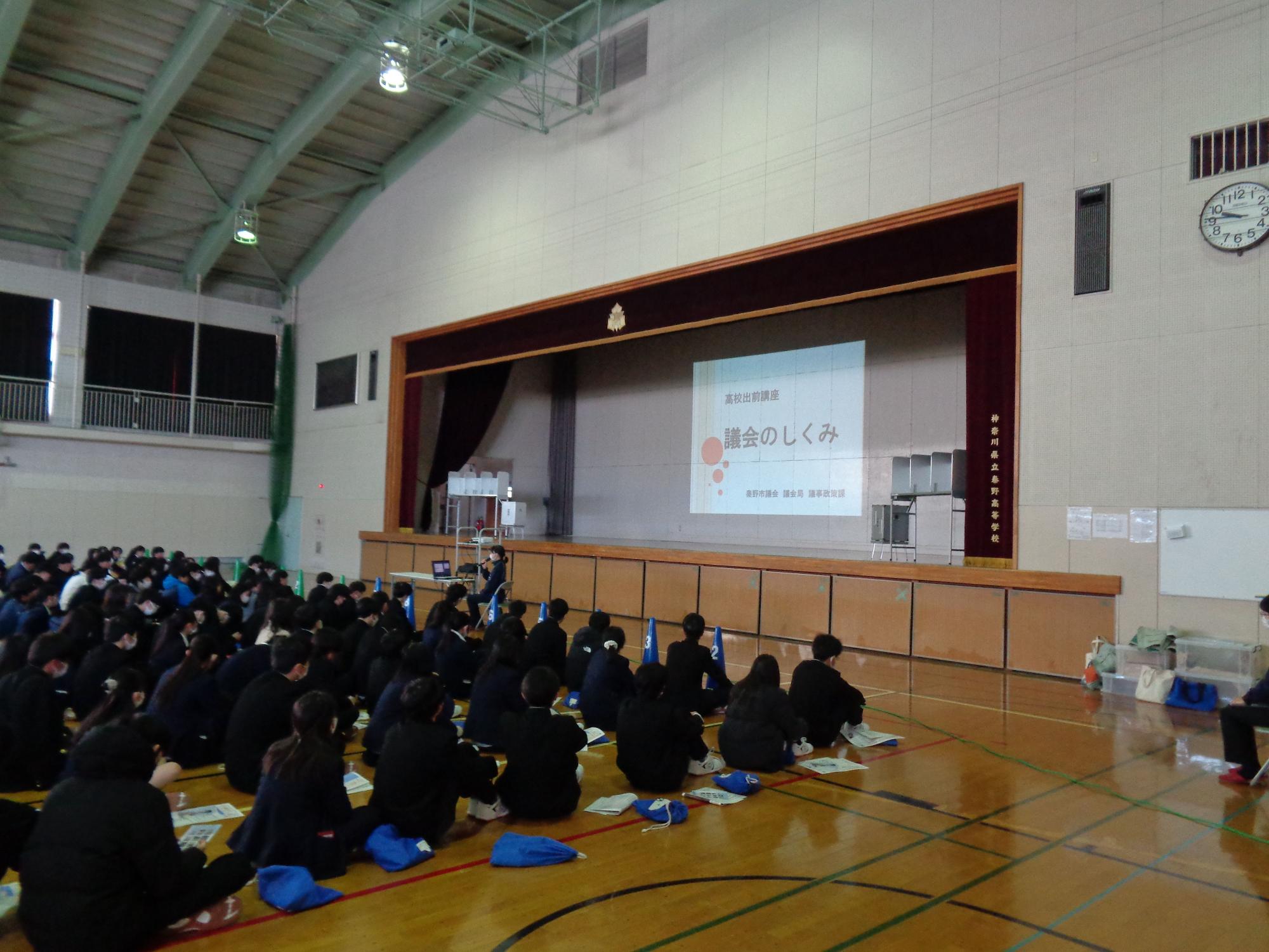 体育館の床に黒い制服姿の生徒が大勢座って壇上を見ており、中央のスクリーンには「議会のしくみ」というタイトルがが投影されている、高校生への出前講座の写真