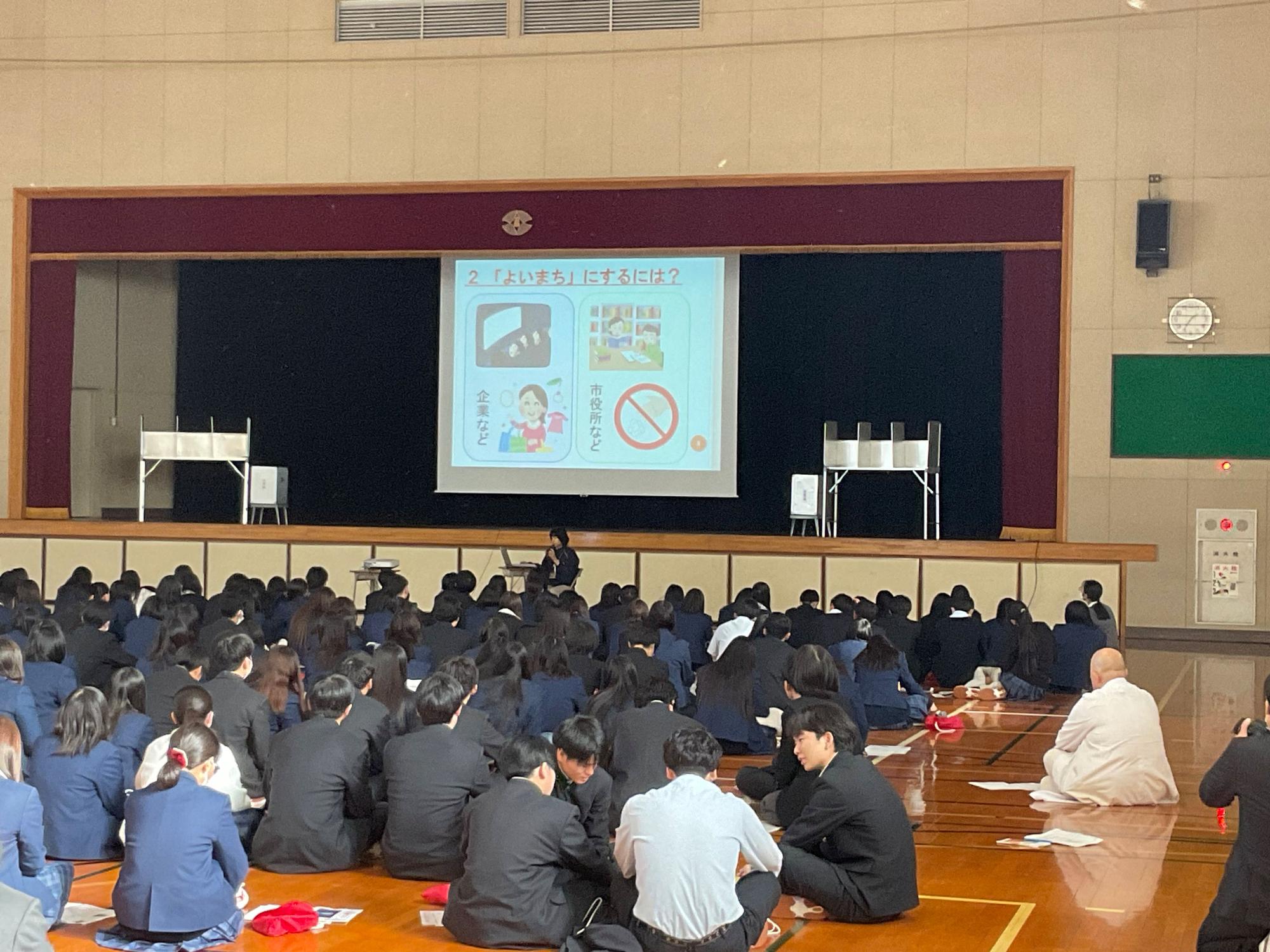大勢の制服姿の生徒が体育館の床に座って壇上を見ており、スクリーンには「2 『よいまち』にするには？」というタイトルや禁止のマークのイラストが描かれたスライドが投影されている出前講座の写真