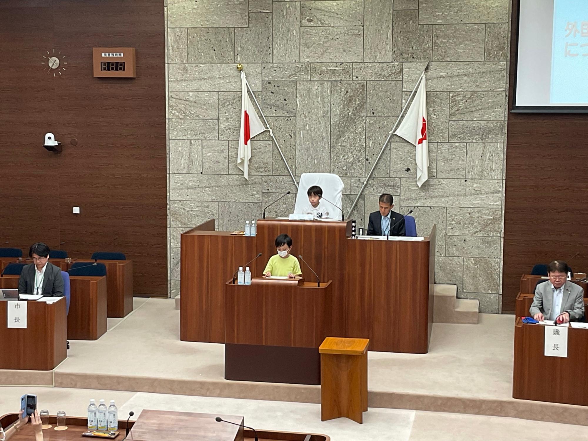 市議会の本会議場で、黄色のTシャツを着た男の子が演壇に立ち、スピーチをしている様子の写真