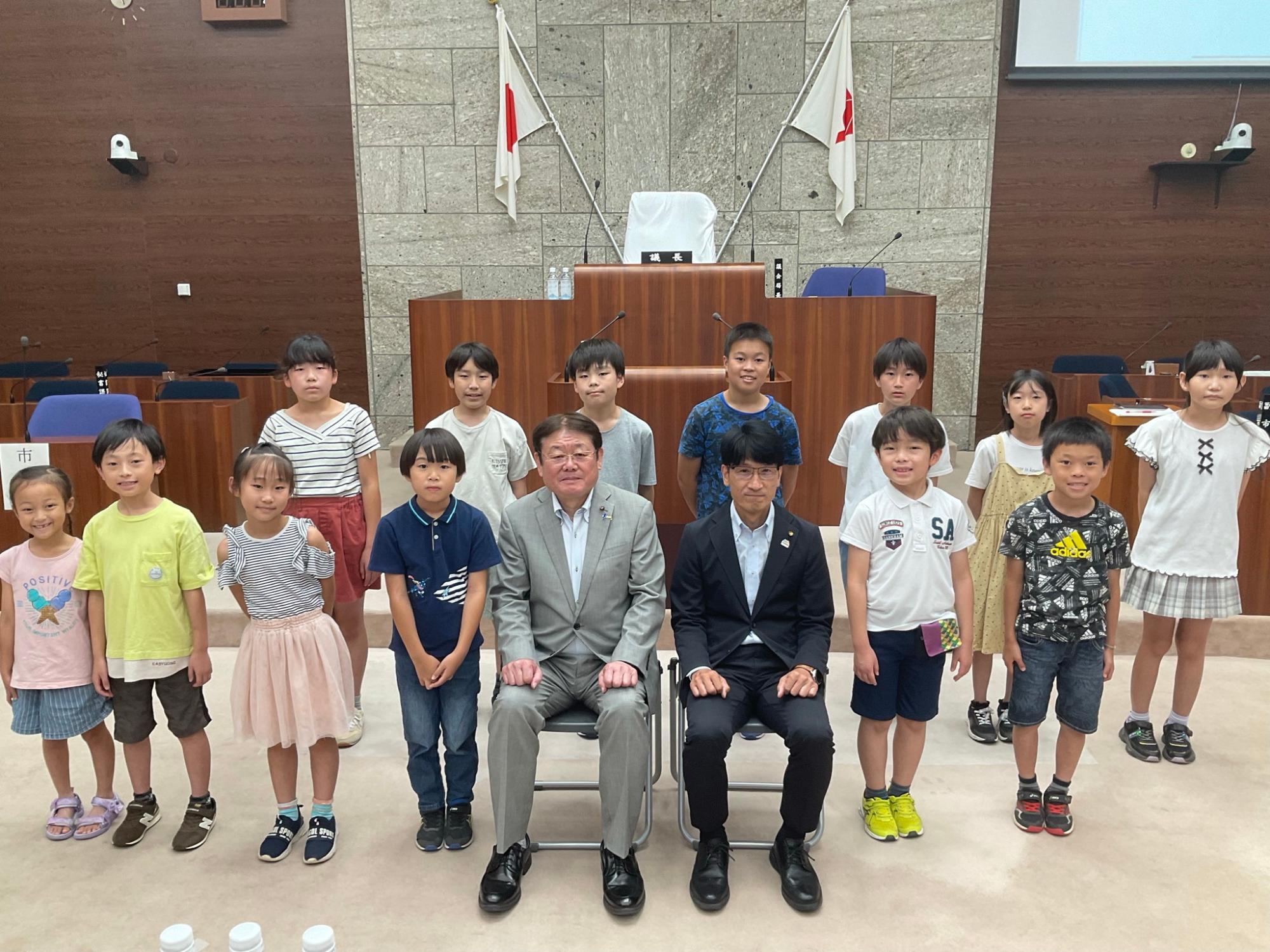 木目調の議場で、日本の国旗を背景に、スーツ姿の大人2人が椅子に座っており、その周りを子供たちが囲んで記念撮影をしている写真