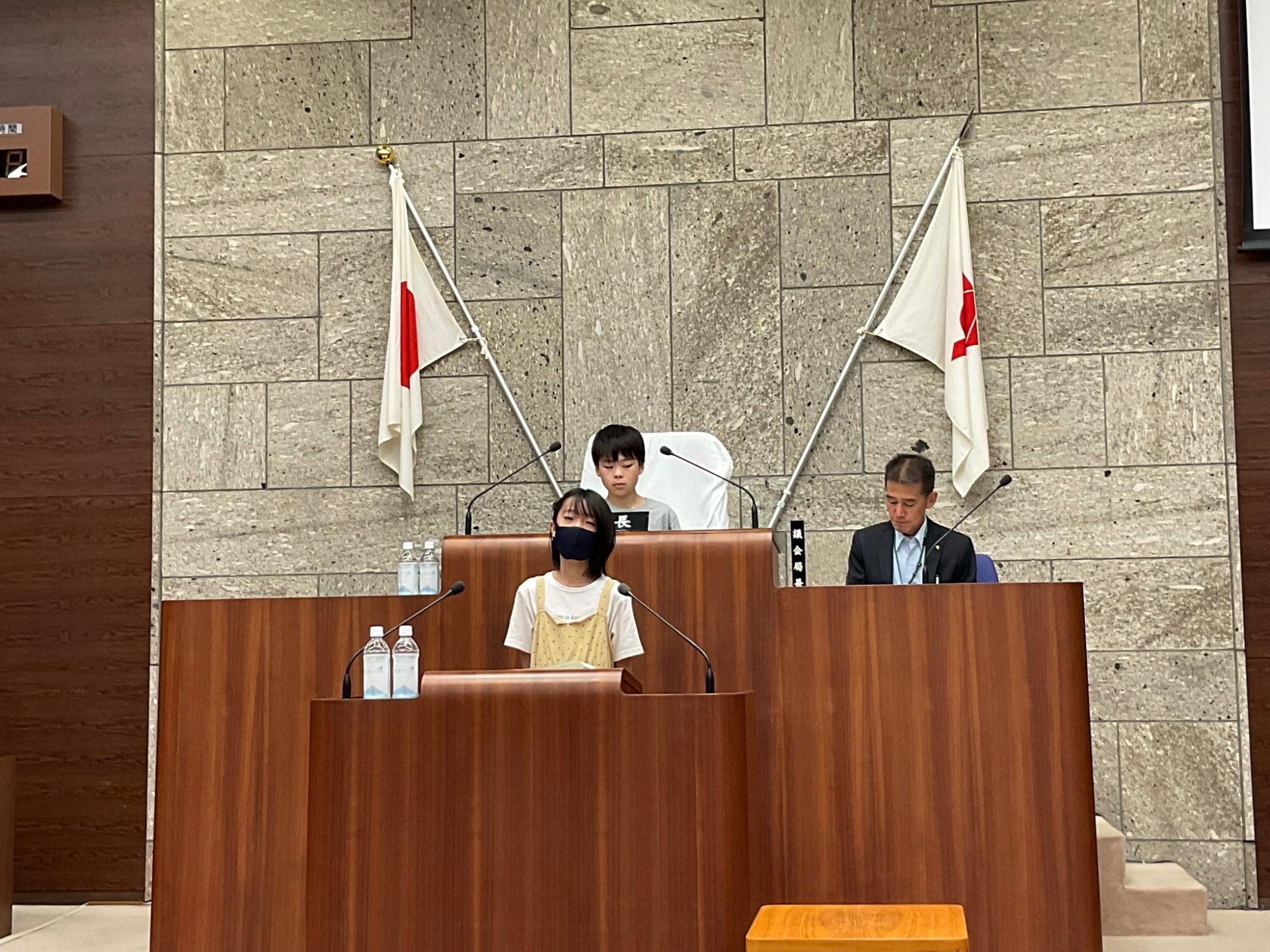 市議会の本会議場で、黒いマスクをした女の子が演壇に立ち、スピーチをしている様子の写真