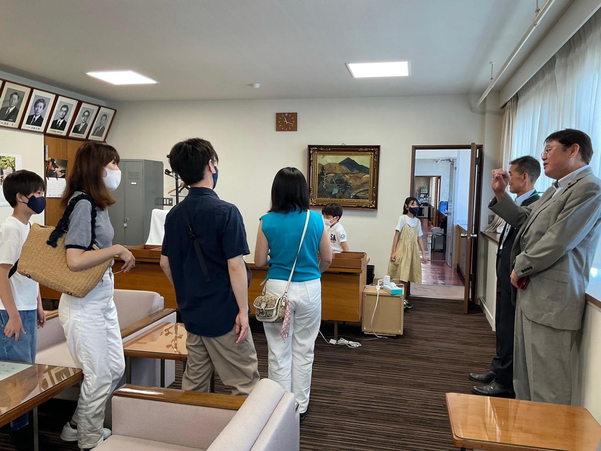 スーツ姿の男性が説明をしており、保護者や子どもたちが興味深そうに耳を傾けている様子の写真