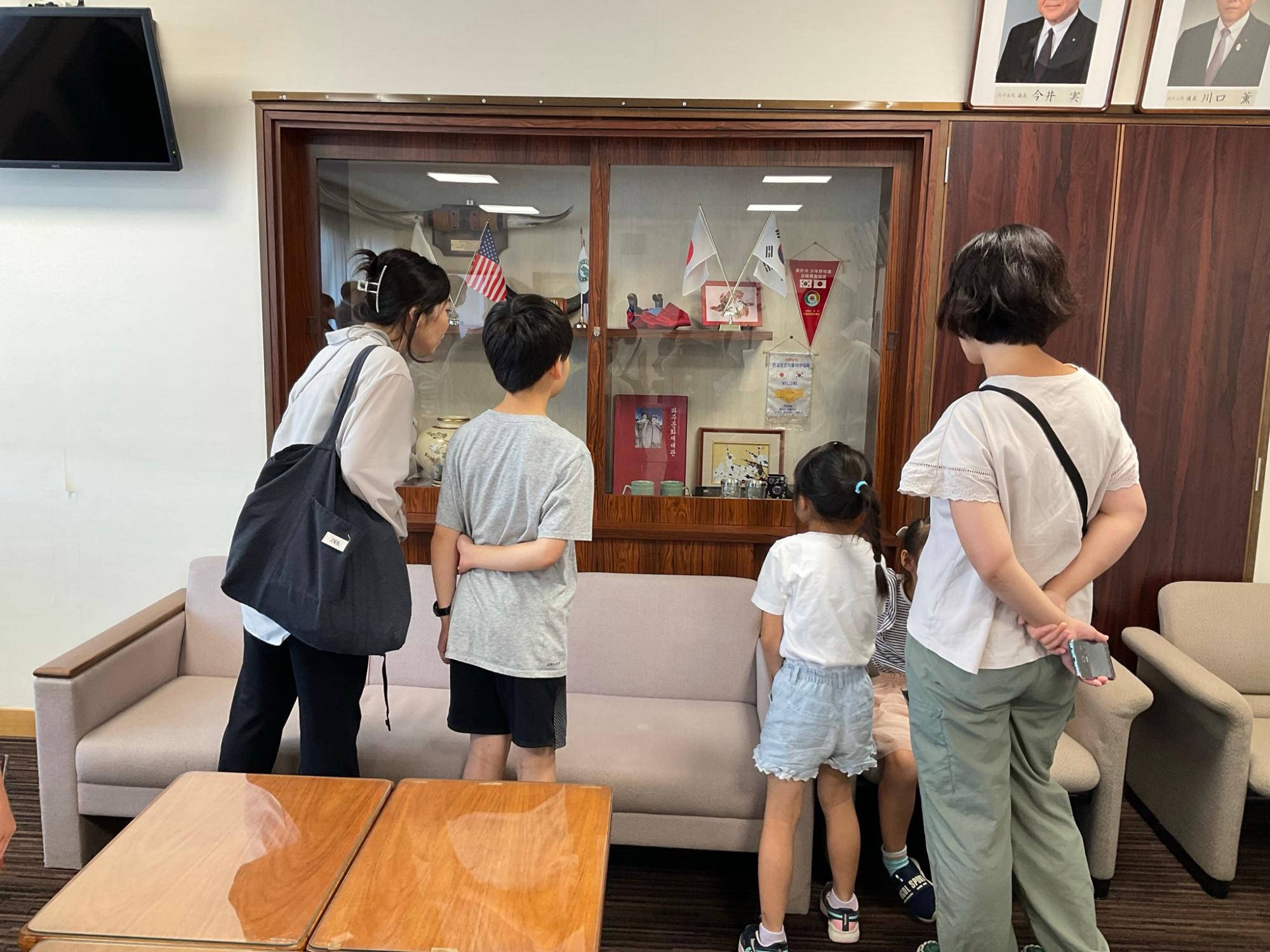 ガラスケースの中に外国の国旗やトロフィーが飾られており、4人が熱心にその展示物を見学している様子の写真