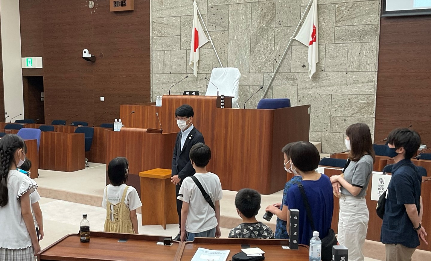 日本の国旗が飾られた議場で、スーツ姿の男性が子供たちに話しかけており、議会見学が行われている様子の写真