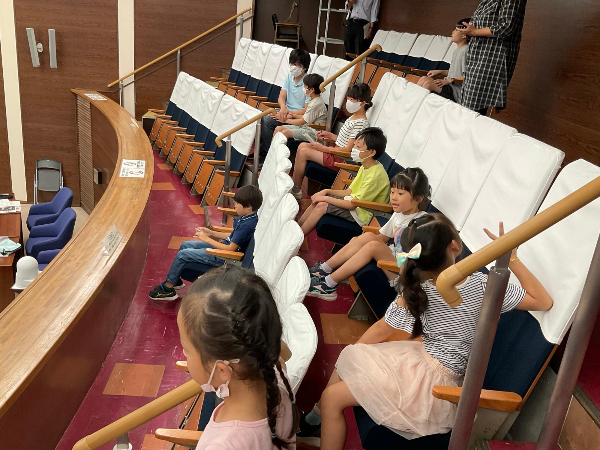 木の机と白い背もたれの椅子が並ぶ議場で、多くの子供たちが着席し、議会見学に訪れている様子の写真