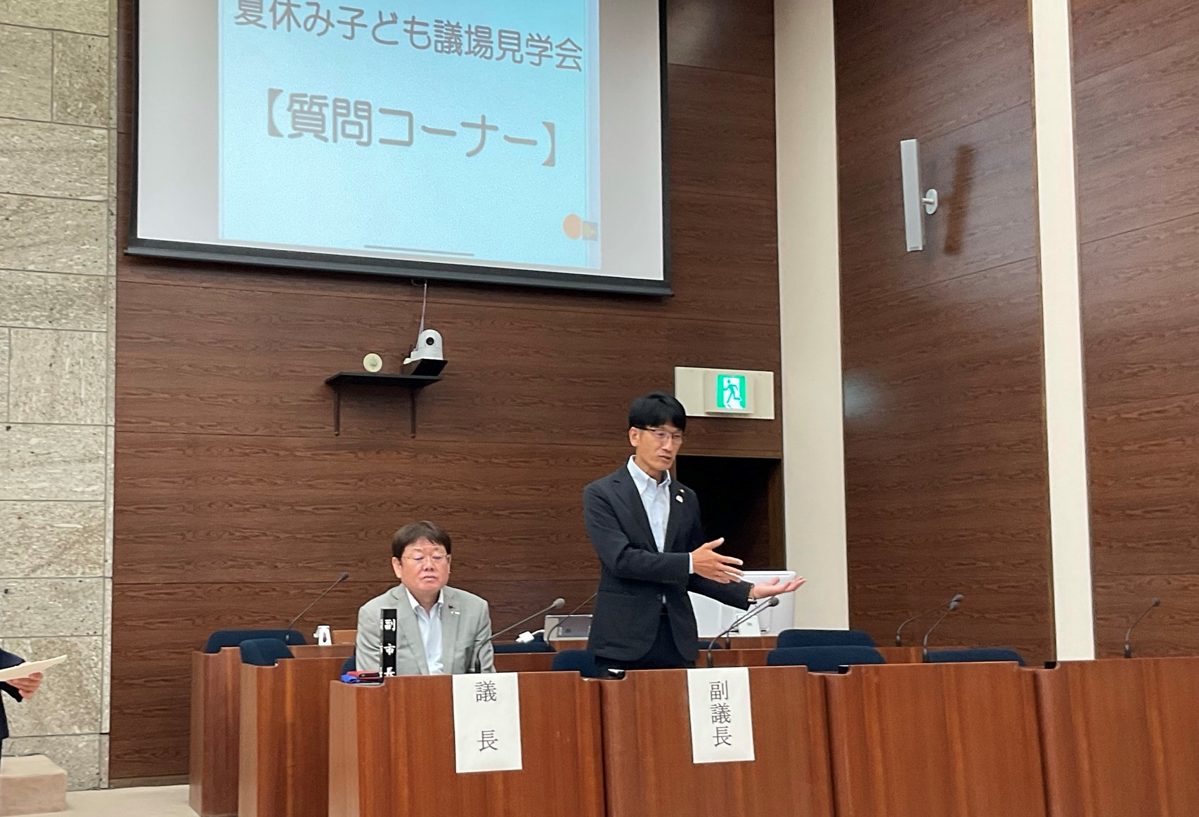 「夏休み子ども議場見学会【質問コーナー】」と書かれたスクリーンを背景に、スーツ姿の男性が議場で話をしている写真