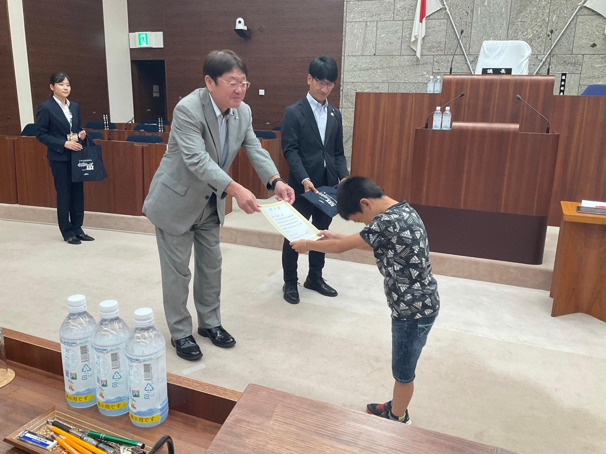 日本の国旗がある議場で、スーツ姿の男性が頭を下げている男の子に修了証を手渡している写真