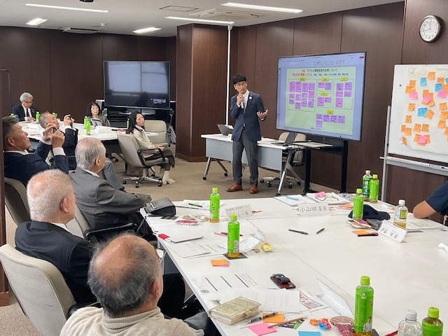 スーツ姿の男性が大型モニターを使って発表し、複数の高齢男性を含む参加者がテーブルを囲んで資料と飲料を前に傾聴している会議室の写真