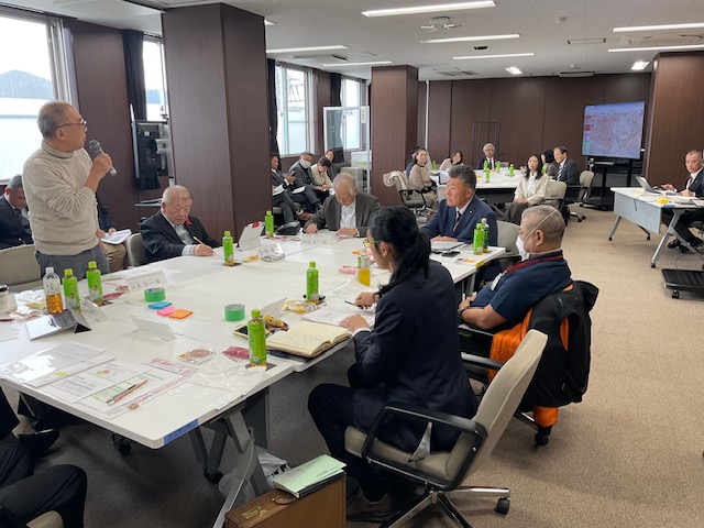 立ってマイクで発言する男性を中心に、複数の参加者が資料と飲料を前に真剣に耳を傾ける会議室内の討議風景の写真