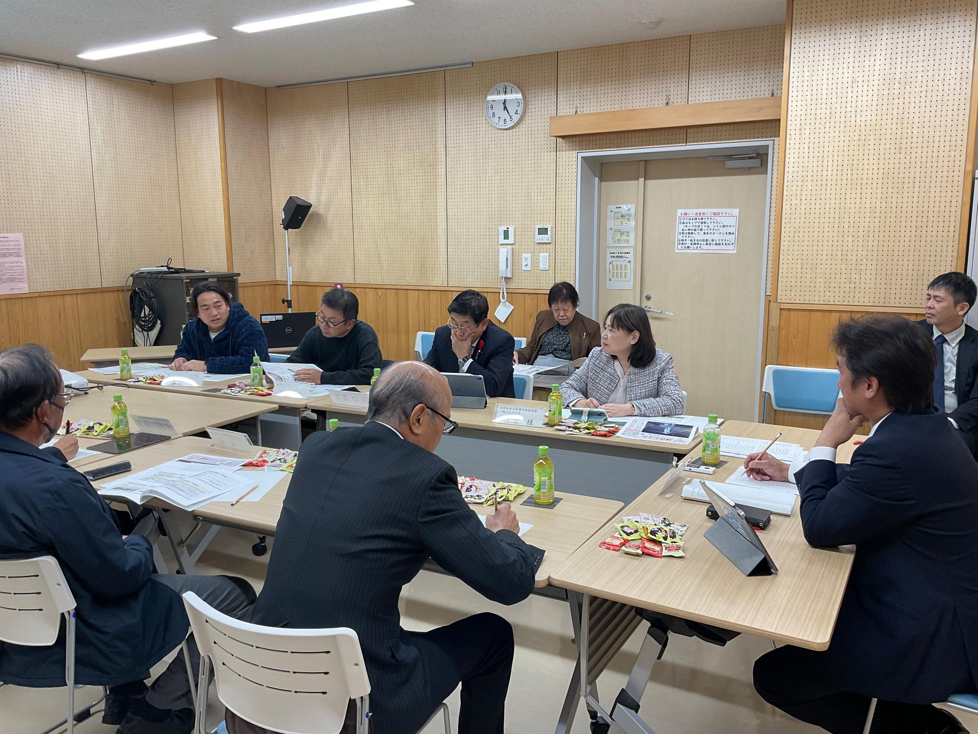 会議室で複数の男女が資料やタブレットを前に円卓状に座り、発言者の話に耳を傾けながら議論を交わしている様子の写真